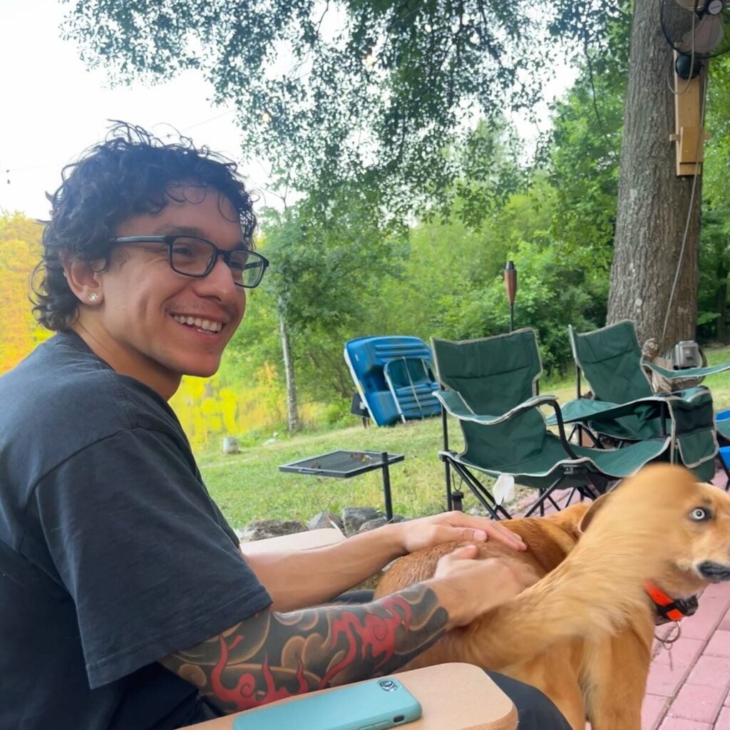 A lab member with glasses and a tattooed arm sits on a wooden chair outdoors, smiling while petting a tan dog. Foldable chairs and trees are visible in the background.