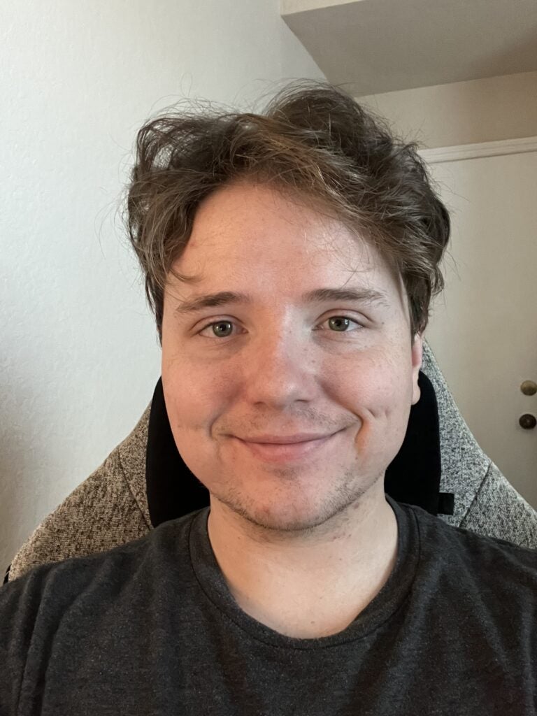 A person with light skin and wavy brown hair, likely one of the lab members, sits in a chair, smiling slightly while looking at the camera. The background shows a white wall and a closed door.
