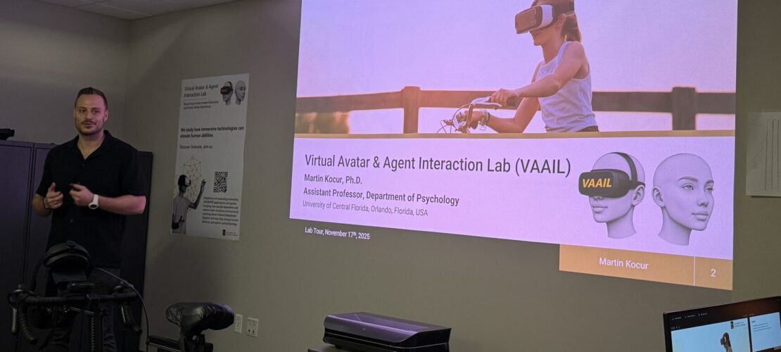 A man stands beside a projected presentation slide about the Virtual Avatar & Agent Interaction Lab at the University of Central Florida, featuring a woman wearing a VR headset.