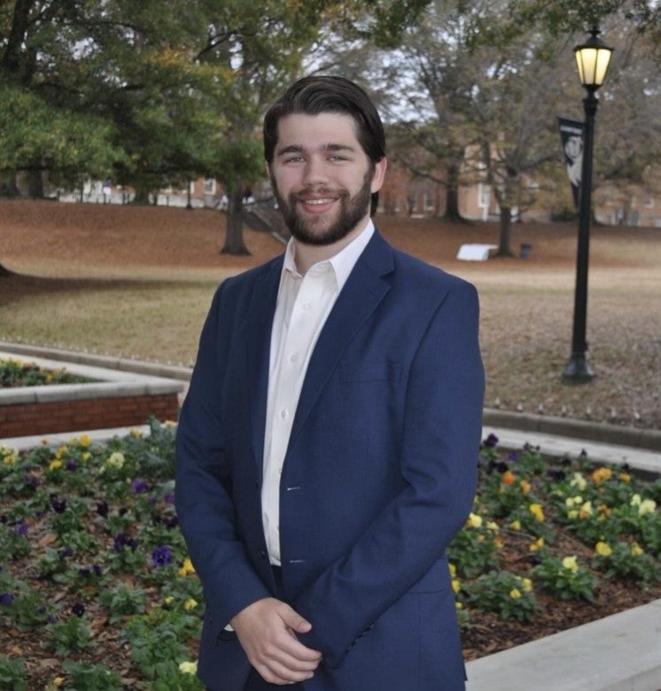 A man in a blue suit stands in front of a flower bed on a college campus, with trees, grass, and a lamppost in the background, appearing ready to meet with fellow lab members.