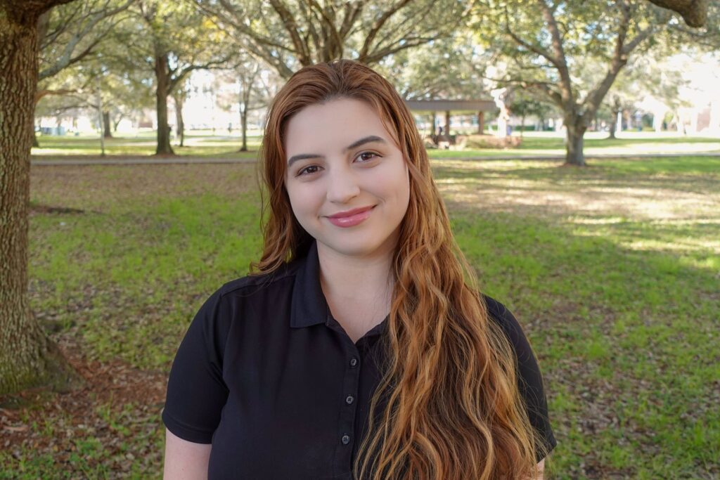 Young woman with long wavy hair wearing a black polo shirt stands outdoors in a park-like setting with trees and grass, appearing as one of the lab members enjoying some time outside.