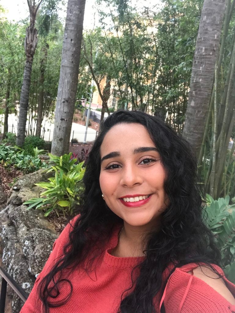 A lab member with long dark wavy hair and red lipstick smiles while posing outdoors.