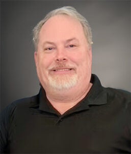 Man with short gray hair and a beard, wearing a black shirt, smiles at the camera against a gray background.