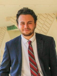 A person with short curly hair and a beard is wearing a dark suit, white shirt, and red striped tie, standing in front of a beige geometric background, smiling at the camera.