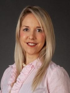 Woman with long blonde hair wearing a light pink blouse smiles at the camera against a dark background.