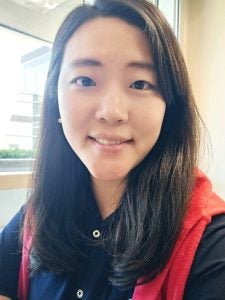 A woman with long dark hair wearing a black shirt and a red sweater is smiling while taking a selfie indoors by a window.