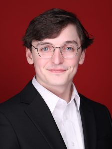 A man with glasses and short brown hair is wearing a black suit jacket, white dress shirt, and is standing against a solid red background.