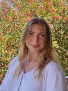 A person with long, blonde hair stands in front of a bush with red flowers, smiling at the camera. The sky is clear and blue in the background.