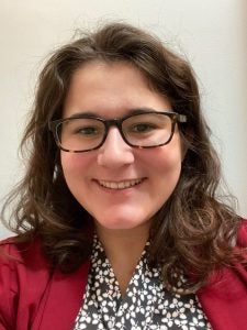 Person with curly brown hair and glasses, wearing a patterned blouse and a red jacket, smiling at the camera against a plain background.