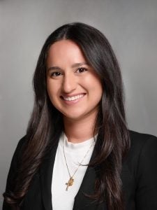 A woman with long dark hair wearing a black blazer and white top is smiling at the camera. She is wearing a cross necklace and has a neutral background behind her.