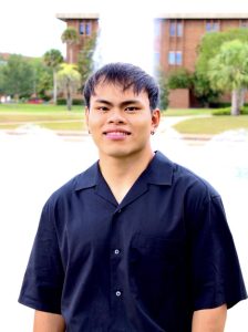 A person stands outdoors in front of a water feature, wearing a dark short-sleeved shirt and light-colored pants, smiling at the camera. Trees and buildings are visible in the background.