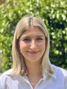 A woman with shoulder-length blonde hair is smiling, standing outdoors in front of a leafy green background, wearing a white blouse and a silver necklace.