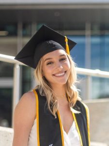 A smiling graduate wearing a black cap and gown stands outside a building.