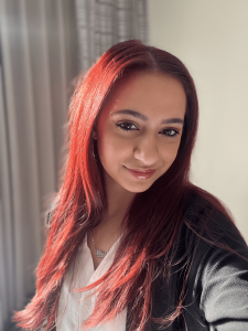 Person with long red hair, wearing a white shirt and black jacket, smiling at the camera in a well-lit room.