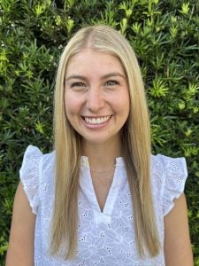 A woman with long blonde hair and wearing a white eyelet blouse stands smiling in front of a dense green shrub.