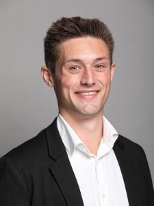 A man with short brown hair, wearing a white shirt and black blazer, smiling in front of a grey background.