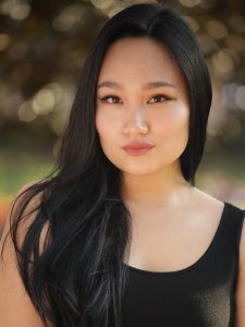 A woman with long black hair and a serious expression is wearing a black tank top, posing outdoors with a blurred background.