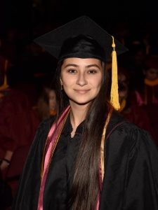 Young woman in a black graduation cap and gown with a gold tassel and maroon cords stands facing the camera with a slight smile.