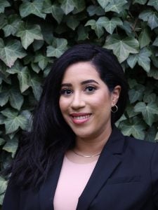 A woman with long dark hair smiles at the camera. She is wearing a black blazer and a light-colored top. The background consists of lush green ivy leaves.