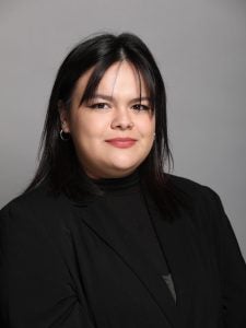 A person with medium-length dark hair wearing a black blazer and black top, standing against a plain grey background.