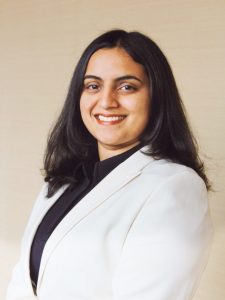 A woman with shoulder-length black hair, wearing a white blazer and black shirt, smiles while looking at the camera.