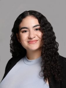 Portrait of a woman with long curly hair, wearing a light blue top and black blazer, smiling against a gray background.