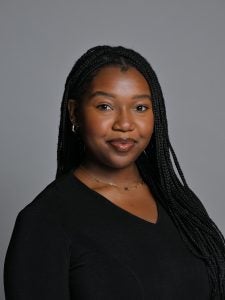 A woman with long braided hair wearing a black top poses for a portrait against a gray background.