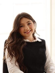 A woman with long, dark hair sits at a table, holding a document and smiling at the camera. She is wearing a white blouse under a black vest.