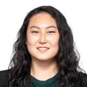 A woman with long, wavy black hair, wearing a green top and black jacket, smiles at the camera against a white background.