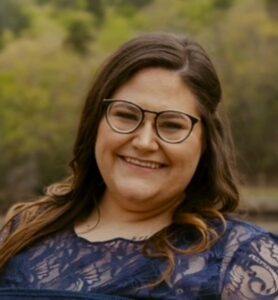 A woman with long brown hair and glasses smiles at the camera, wearing a navy lace top, with greenery blurred in the background.