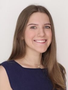 A young woman with straight brown hair, wearing a navy sleeveless top and a small necklace, smiles in front of a plain white background.