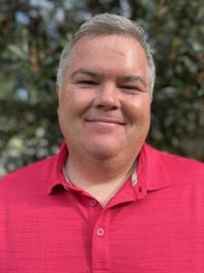 A man with short gray hair wearing a bright pink collared shirt stands outdoors, smiling at the camera with greenery in the blurred background.