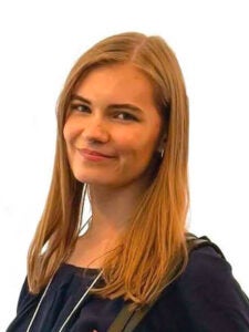 A young woman with straight, light brown hair is wearing a dark top and backpack, smiling slightly, photographed against a plain white background.