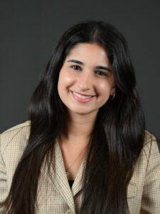 A woman with long dark hair, wearing a light plaid blazer, smiles at the camera in front of a plain dark background.