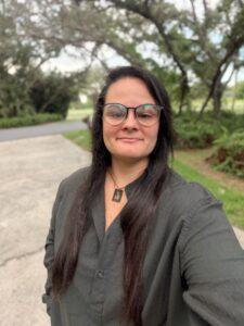 A person with long dark hair and glasses, wearing a dark button-up shirt and necklace, stands outdoors on a concrete path with trees and greenery in the background.