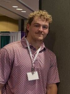 A person wearing a patterned pink shirt and name badge stands indoors near a partition at what appears to be an event or conference.