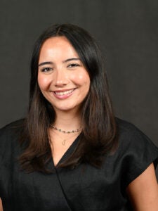 Woman with long dark hair wearing a black top, a necklace, and smiling, posed in front of a plain dark background.