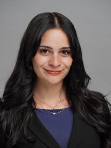 Woman with long dark hair wearing a black blazer and blue top, smiling at the camera against a plain gray background.