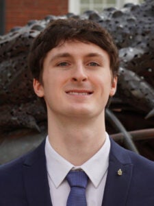 Young man in a navy suit and tie standing outside in front of a textured metal sculpture, smiling at the camera.