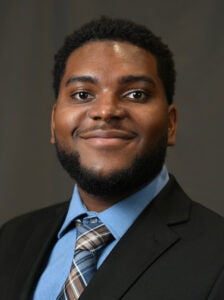A man wearing a dark suit, blue shirt, and patterned tie poses in front of a plain dark background, looking at the camera and smiling slightly.