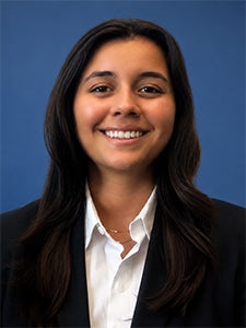 Woman with long dark hair wearing a black blazer and white shirt, smiling at the camera, in front of a plain blue background.