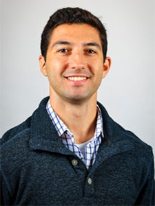A man with short dark hair, wearing a blue pullover sweater over a checkered shirt, smiles at the camera against a plain light background.