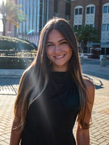Woman with long brown hair wearing a sleeveless black top, standing and smiling outdoors in a sunlit urban area with palm trees and buildings.