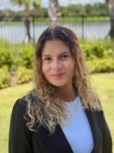 A woman with curly hair wearing a black blazer and white top stands outside on grass with palm trees and a lake in the background.
