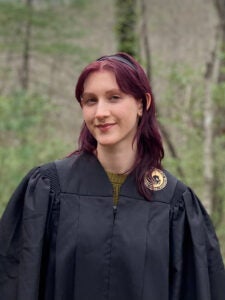 A person with reddish hair wearing a black graduation gown stands outdoors in front of trees and greenery.