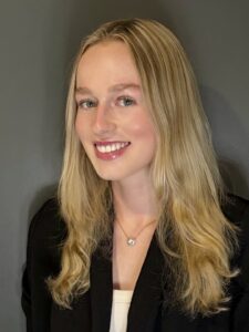 Young woman with long blonde hair, wearing a black blazer and white top, smiling in front of a plain dark gray background.