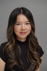 A woman with long wavy brown hair wearing a black top is facing the camera against a plain gray background.