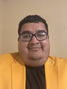 A person with short dark hair, glasses, and a trimmed beard is wearing a yellow blazer and black shirt, posing in front of a plain beige wall.