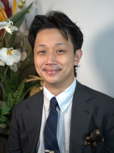 A person in a suit and tie sits in front of a plant arrangement, facing the camera and smiling slightly.