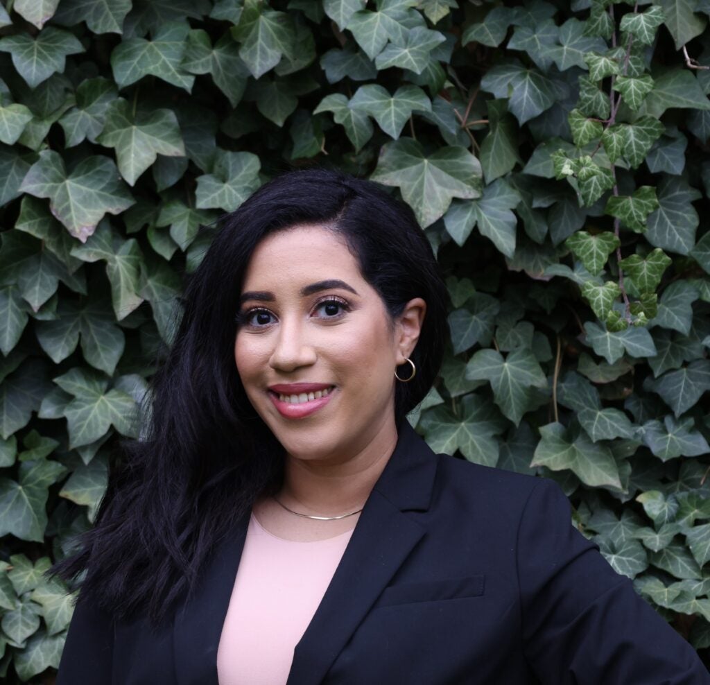 Woman with long dark hair wearing a black blazer and light top, standing in front of a wall covered with green ivy leaves.
